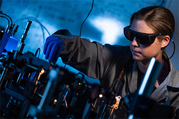 A fair-skinned woman with brunette hair is touching optics with a gloved hand while wearing laser safety goggles.